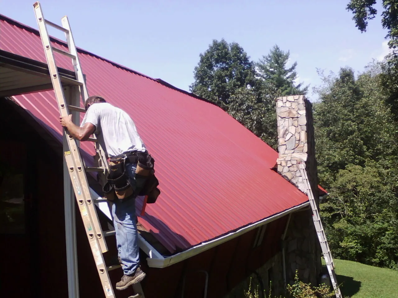Roofer on a ladder beside a red metal roof and stone chimney — Soffit & Fascia Repair by Roofing Brothers in Zeigler